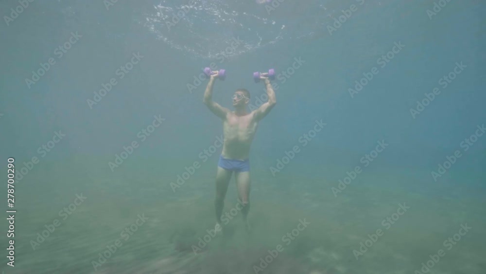 Athletic man does sport exercise with dumbbells underwater stands on ocean floor. Athletic man does sport exercise with dumbbells underwater stands on ocean floor.