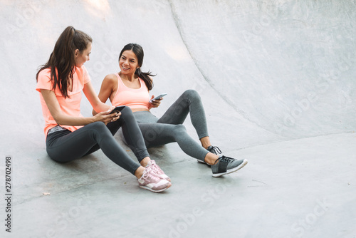 Image of happy fitness women in sportswear holding cellphones together while sitting on concrete sports ground