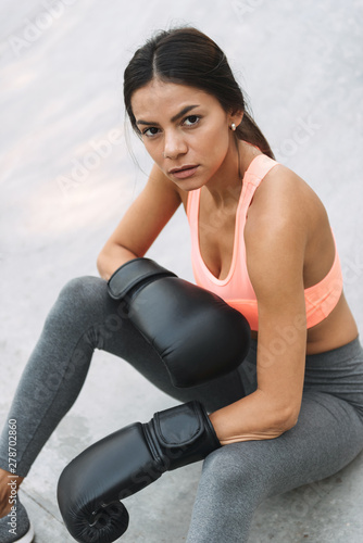 Image of concentrated young woman in boxing gloves sitting on concrete floor outdoors
