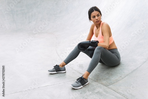 Image of serious young girl in boxing gloves sitting on concrete floor outdoors
