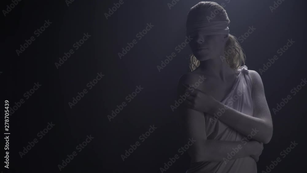 Living statue of Themis with the blindfold on her face is posing in low lighting Living statue of Themis with the blindfold on her face is posing in low lighting