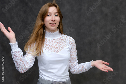 Portrait to the waist of a young pretty brunette girl woman with beautiful long hair on a gray background in a white jacket. He talks, smiles, shows his hands with emotions in various poses.