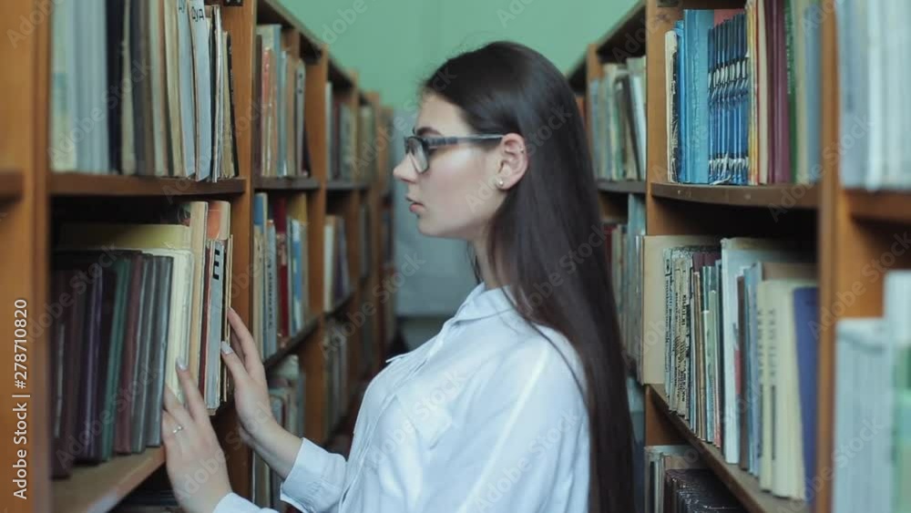 Smart looking and beautiful young girl in white shirt and glasses in collage school library selecting book to read Smart looking and beautiful young girl in white shirt and glasses in collage school library selecting book to read