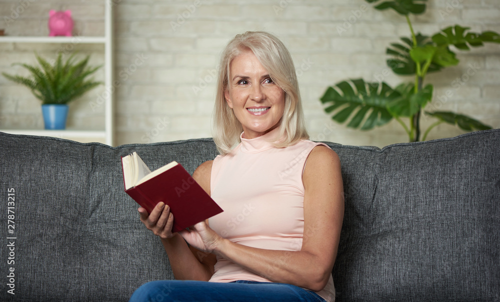 Happy middle aged woman is reading a book at home - obrazy, fototapety, plakaty 