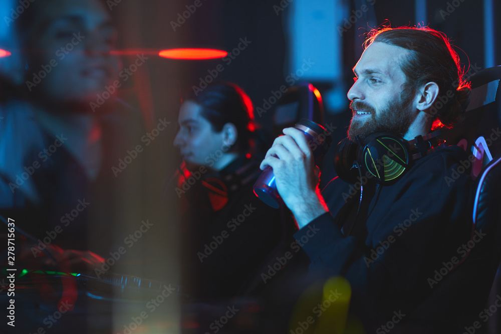 Smiling satisfied young bearded gamer with headphones on neck sitting in comfortable armchair and drinking energy beverage while playing video game