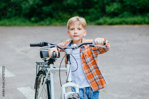Child boy in orange shirt carries a big bike in the summer park outdoor.