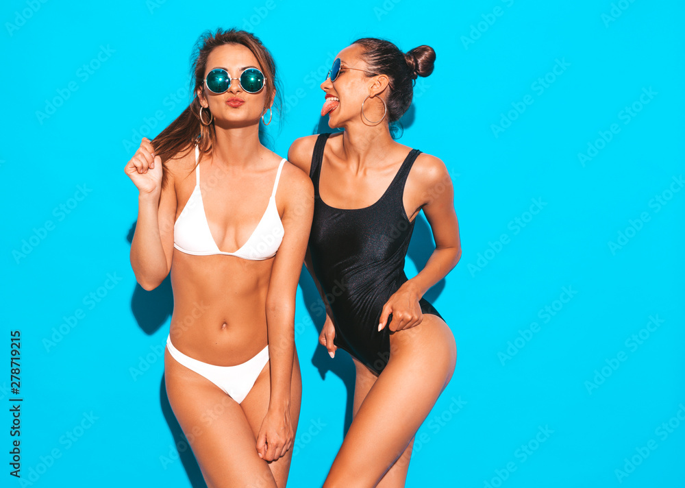 Portrait of two beautiful sexy smiling hipster women in summer white and black swimwear bathing suits. Trendy hot models having fun in studio. Girls isolated on blue.Playing with hair in sunglasses - obrazy, fototapety, plakaty 
