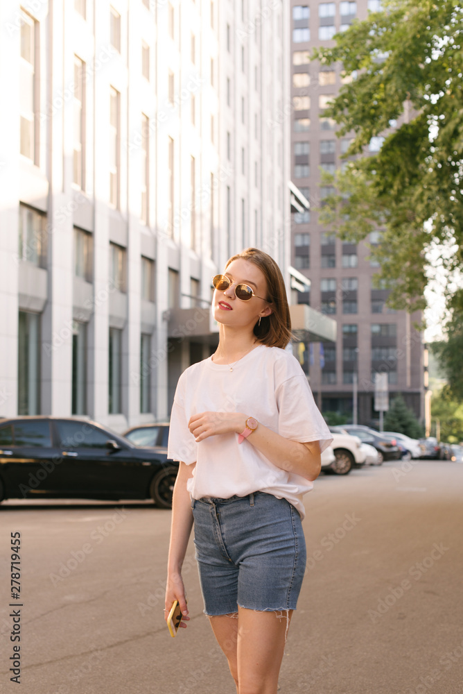 Street portrait of a stylish girl in casual clothing for a walk on a warm summer day. Attractive girl in the sunglasses poses on the street in the background of the urban landscape. Vertical photo.