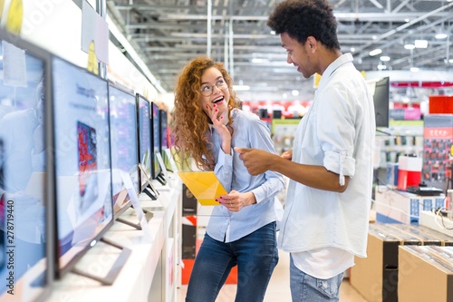 African guy with red-haired girl standing in front of the stand in the electronics store choose a plasma TV looking at the price tags