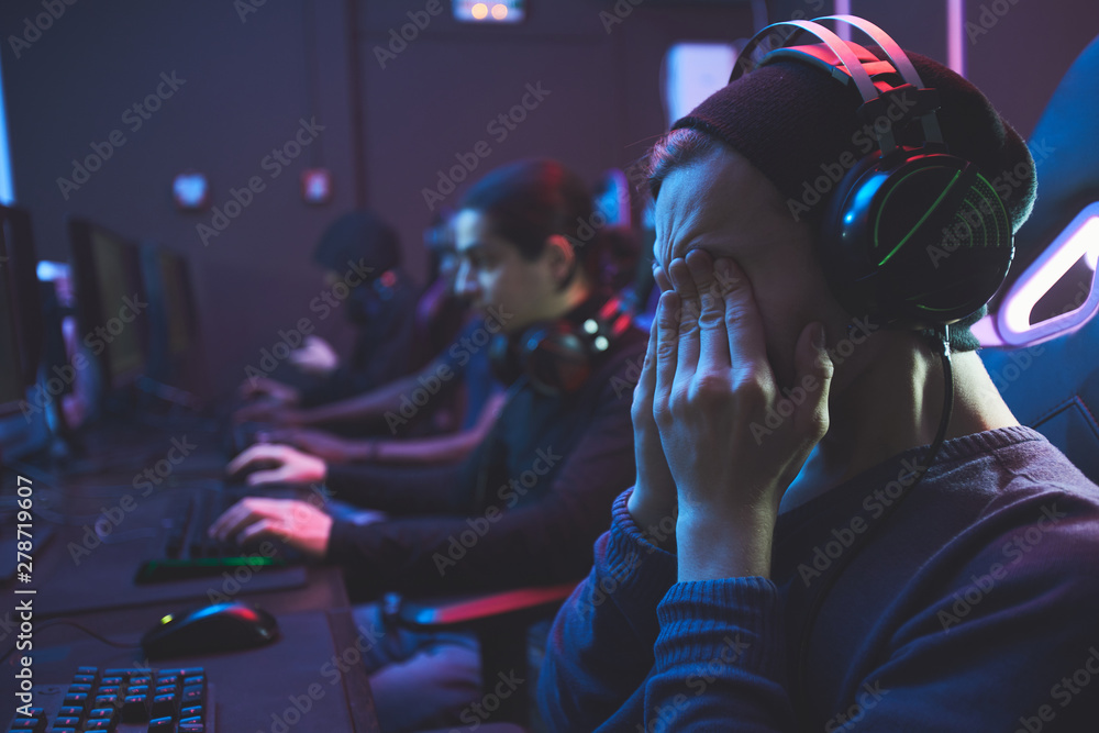 Young geek guy in headset rubbing eyes while feeling eye fatigue from computer monitor, he playing video game Young geek guy in headset rubbing eyes while feeling eye fatigue from computer monitor, he playing video game