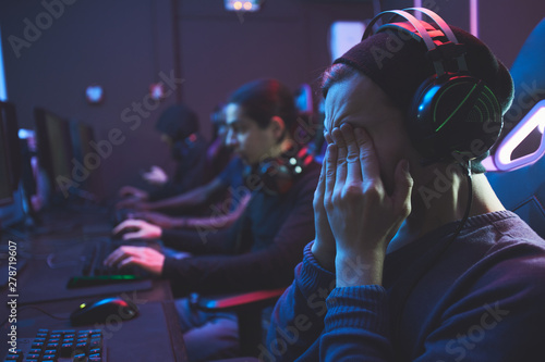 Young geek guy in headset rubbing eyes while feeling eye fatigue from computer monitor, he playing video game photo