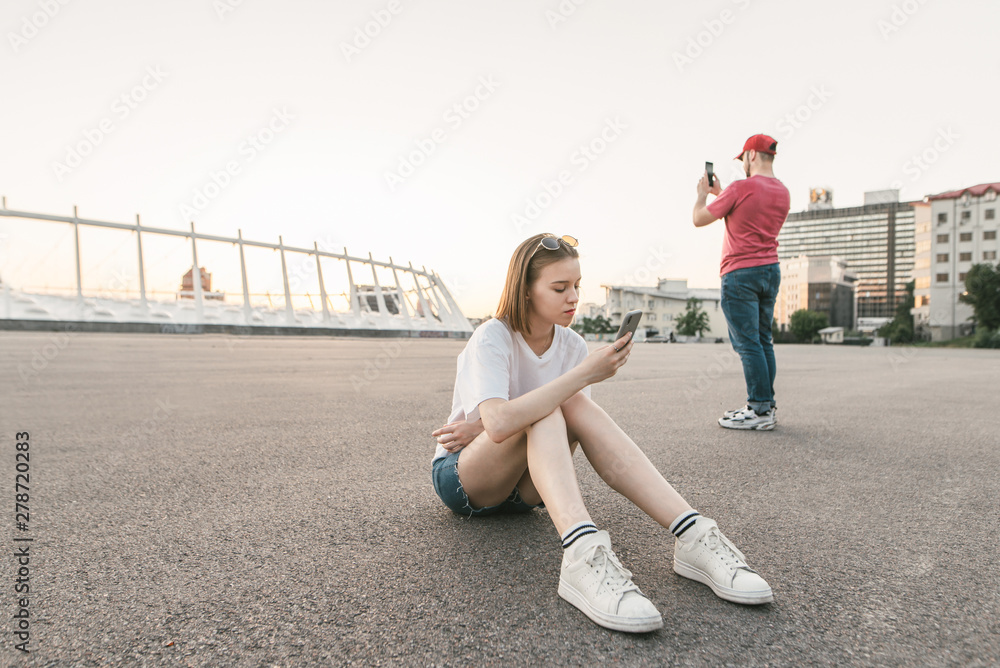 Fototapeta premium People use smartphones in the outdoors against the backdrop of urban scenery. The girl uses a smartphone, a man makes a photo. Millennials on a walk with gadgets in their hands.