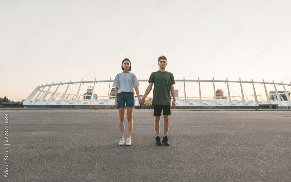 Stylish couple holding hands standing on the background of an evening city and looking in camera. Portrait of the full height of a beautiful guy and a girl on the background of modern architecture