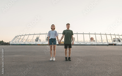 Stylish couple holding hands standing on the background of an evening city and looking in camera. Portrait of the full height of a beautiful guy and a girl on the background of modern architecture