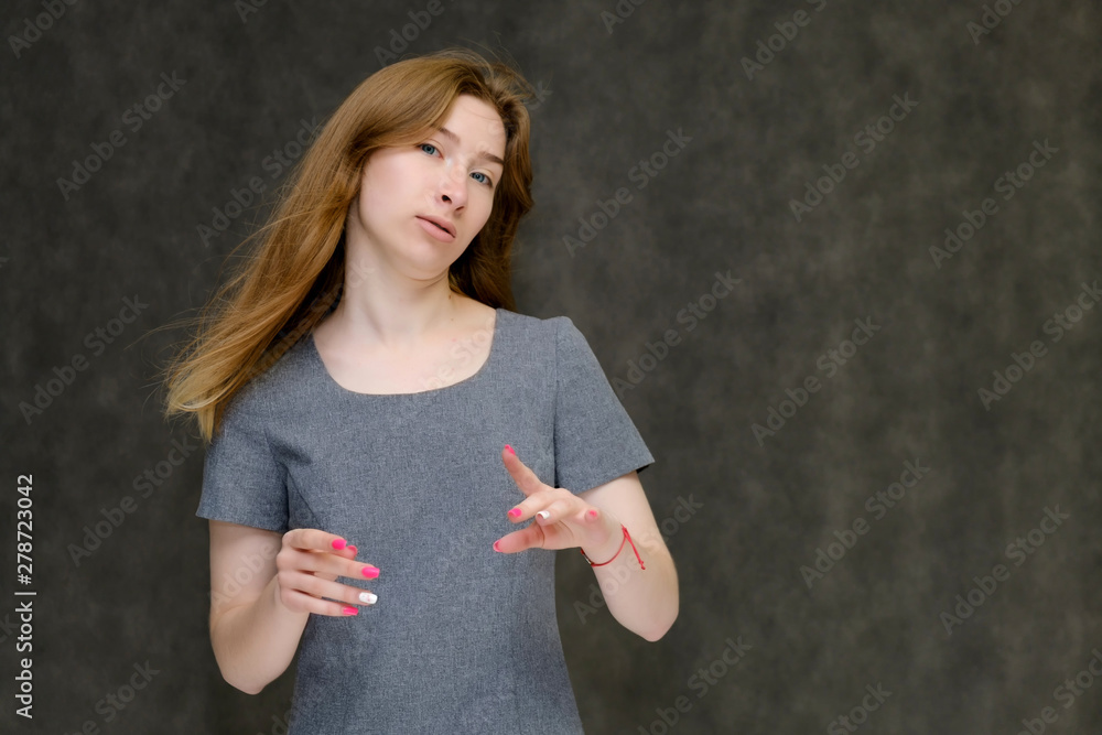 Naklejka premium Portrait to the waist of a young pretty brunette girl woman with beautiful long hair on a gray background in a gray dress. He talks, smiles, shows his hands with emotions in various poses.
