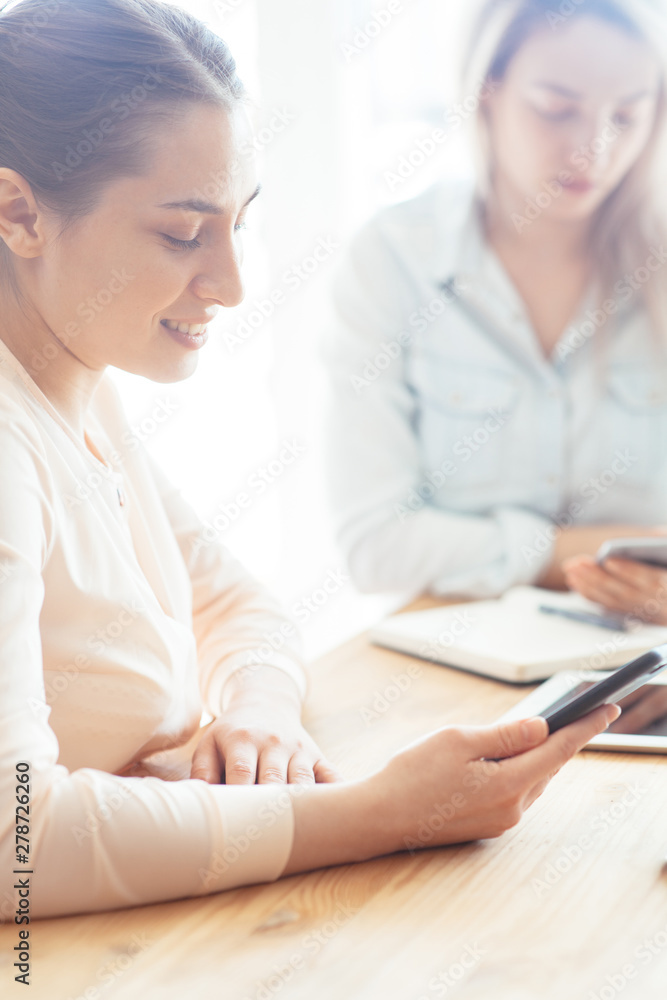 The girl at a business meeting with another girl looks into the phone and smiles The girl at a business meeting with another girl looks into the phone and smiles