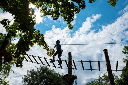 Young boy walking on a rope trail framed by green foliage with cloudy blue sky in the background