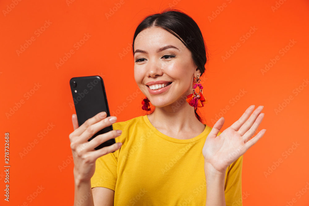 Happy young woman posing isolated over orange wall background talking by mobile phone take a selfie waving. Happy young woman posing isolated over orange wall background talking by mobile phone take a selfie waving.