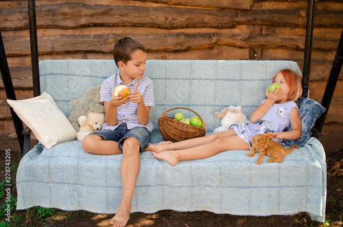 Brother and sister in the garden on the garden swing eating apples. Happy carefree childhood in the village. Smiling children with a red kitten