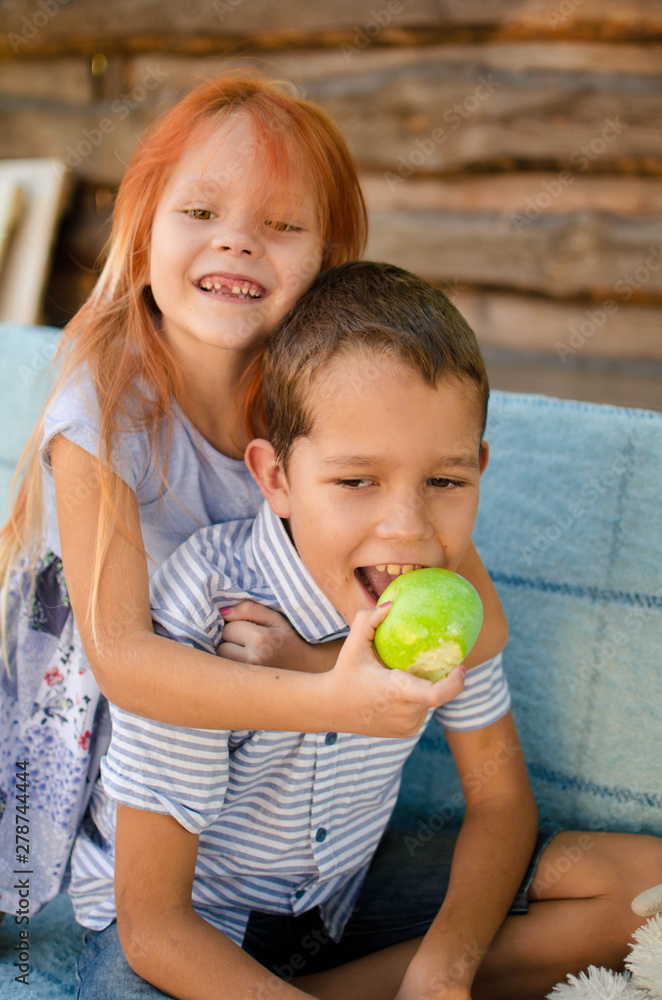 Fototapeta premium Happy boy and girl on a swing in the garden. Sister feeds her brother green apples. Laughing children, happy childhood