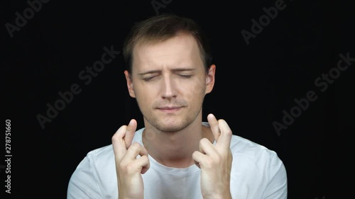 Close up shoot of young attractive caucasian man having his fingers crossed and begging hopefully looking at camera with background isolated on black. Crossed fingers symbolizing good luck.