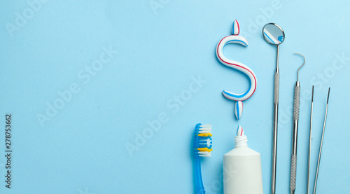 Dollar sign of toothpaste. Tube of colored toothpaste and a toothbrush and dentist tools, a mirror, a hook on a blue background. Concept expensive dentist services. Copy space for text.