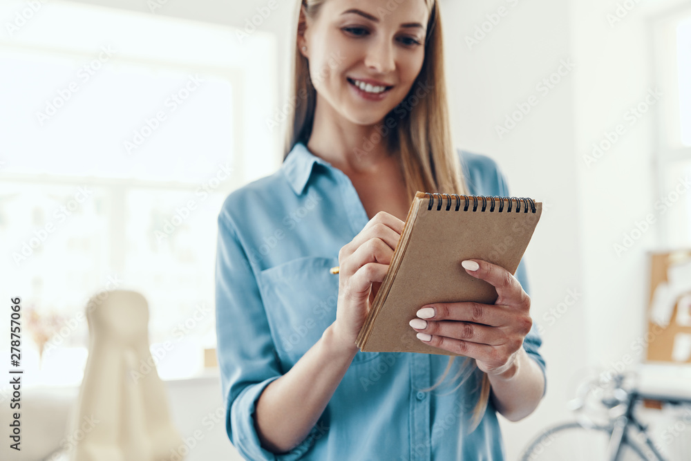 Beautiful young woman in smart casual wear smiling and writing something down while standing in the office