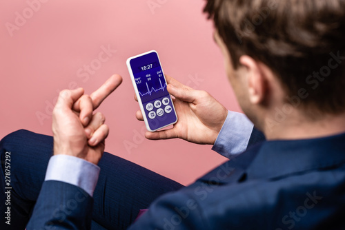 cropped view of businessman using smartphone with health app while showing middle finger, isolated on pink