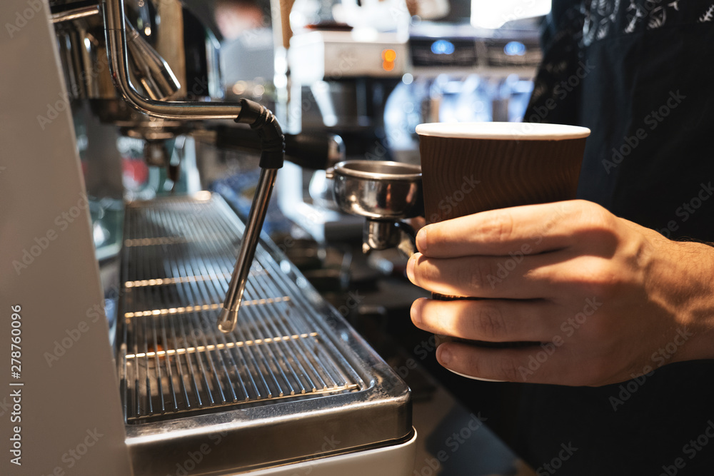 barista man holding hot coffee drink in paper cup in one hand and coffee holder in another standing near professional coffee machine in cafe close up