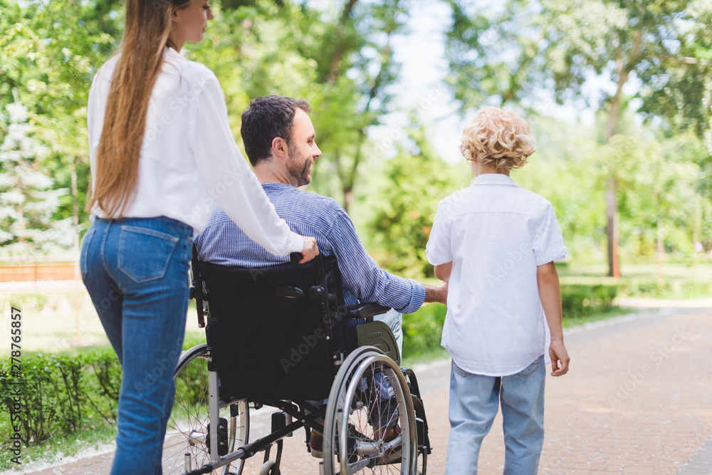 back view of mother rolling wheelchair with disabled father in park back view of mother rolling wheelchair with disabled father in park