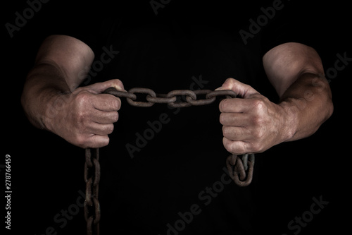 adult man in black clothes stands upright with strained muscles and holds a rusty metal chain