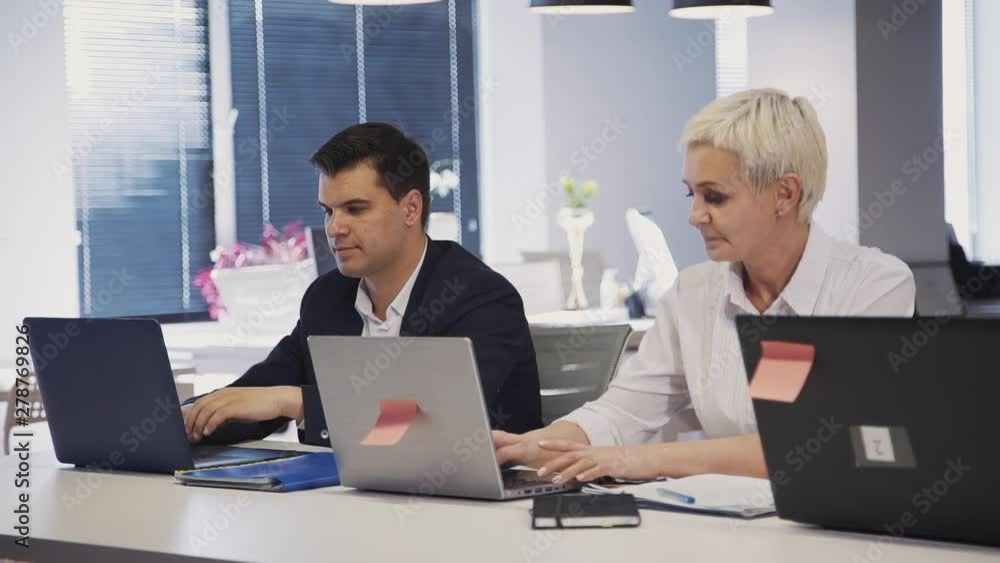 Two mature, qualified and professional colleagues sitting indoor modern office with loft interior work place. Woman and man spending working day using laptop computer and testing program Two mature, qualified and professional colleagues sitting indoor modern office with loft interior work place. Woman and man spending working day using laptop computer and testing program