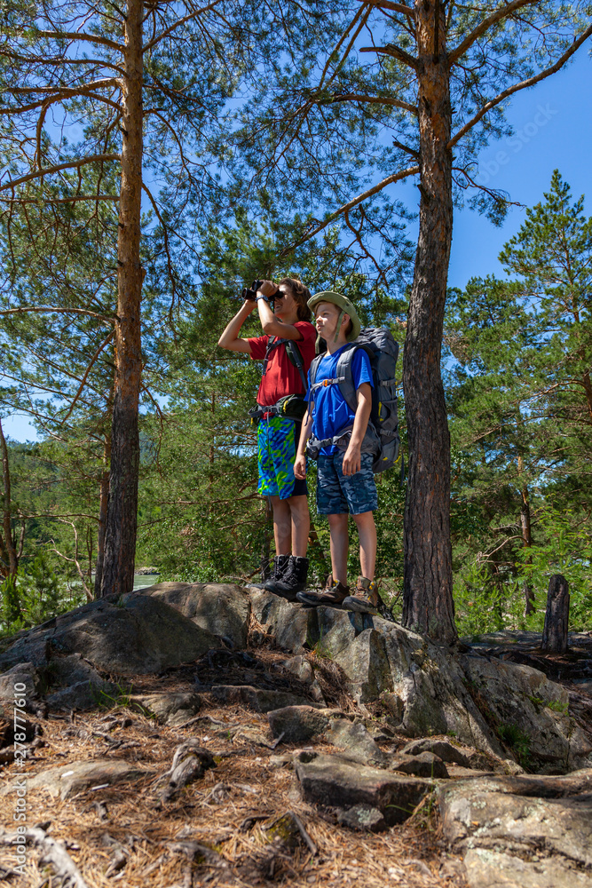 Two boys with backpacks looking through binoculars in the forest. Concept of kids vacations and travel.