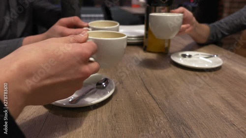 Three friends are sitting at table in cafe. They raised white cups of tea and clink with each other, closeup hands. Group of people meeting and have a rest in evening. photo