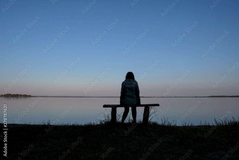 Silhouette of a meditating woman 59 years old at dawn on Svityaz Lake in Ukraine. The woman is sitting on a home-made wooden bench. Self-isolation concept.