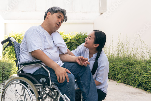 The wife is taking care of the husband sitting on wheelchair,  who is suffering from neurological diseases, or hemiplegia having a facial palsy and kinking fingers, to health and paralysis concept.