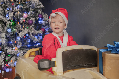 Happy child are dressed in festive costume of Santa Clausn, carrying decorated Christmas tree with balls and garlands on yellow car. Christmas Eve photo