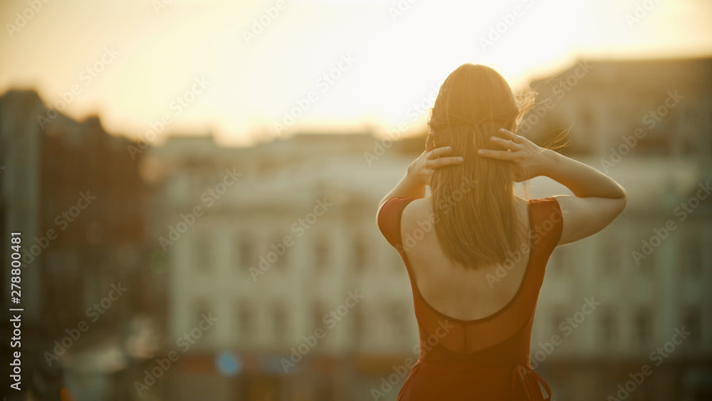 A young woman in red dress standing on the roof and fixing her hair - sunset