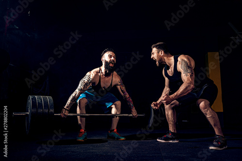 Two muscular bearded tattoed athletes training, one lift heavy weight bar when other is motivating. Scream. Working hard. Exercise for the muscles of the back