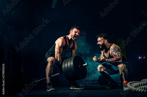 Canvas Print Two muscular bearded tattoed athletes training, one lift heavy weight bar when other is motivating