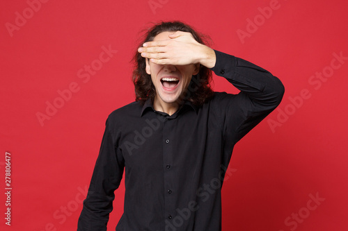 Handsome stylish young curly long haired man in black shirt posing isolated on red wall background studio portrait. People sincere emotions lifestyle concept. Mock up copy space. Cover eyes with hand. © ViDi Studio
