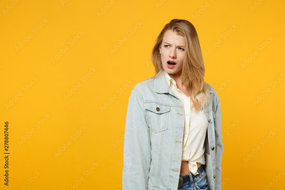 Fototapeta premium Portrait of shocked perplexed young woman in denim casual clothes standing and looking aside isolated on bright yellow orange wall background in studio. People lifestyle concept. Mock up copy space.