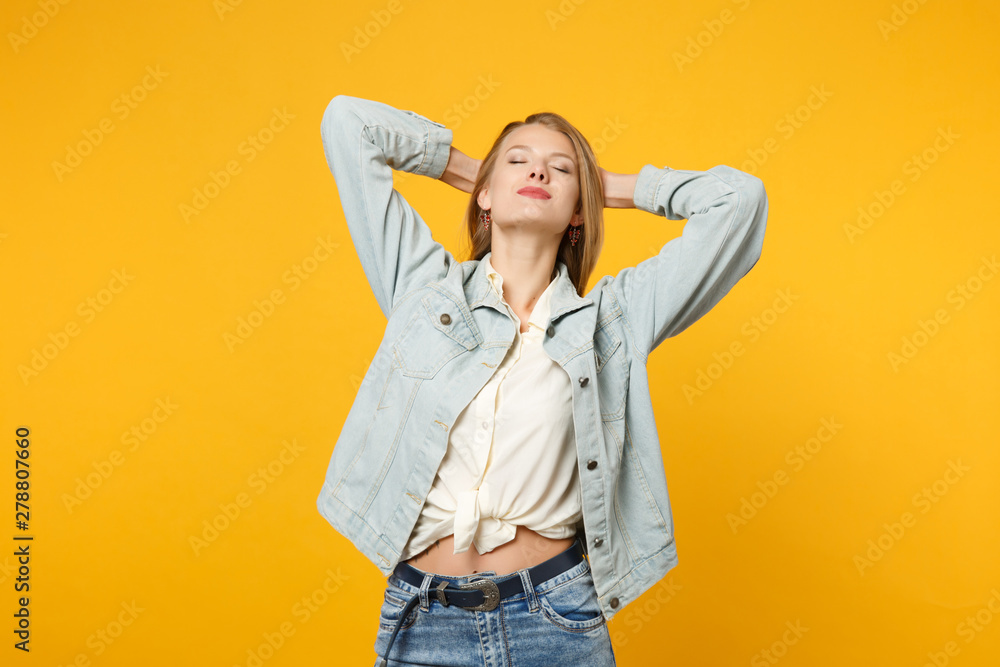 Portrait of relaxed young woman in denim casual clothes keeping eyes closed, holding hands behind her head isolated on yellow orange background in studio. People lifestyle concept. Mock up copy space. Portrait of relaxed young woman in denim casual clothes keeping eyes closed, holding hands behind her head isolated on yellow orange background in studio. People lifestyle concept. Mock up copy space.