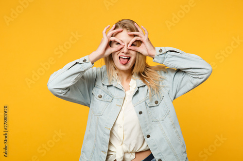 Funny young woman in denim casual clothes hold hands near eyes, imitating glasses or binoculars, showing tongue isolated on yellow orange wall background. People lifestyle concept. Mock up copy space.