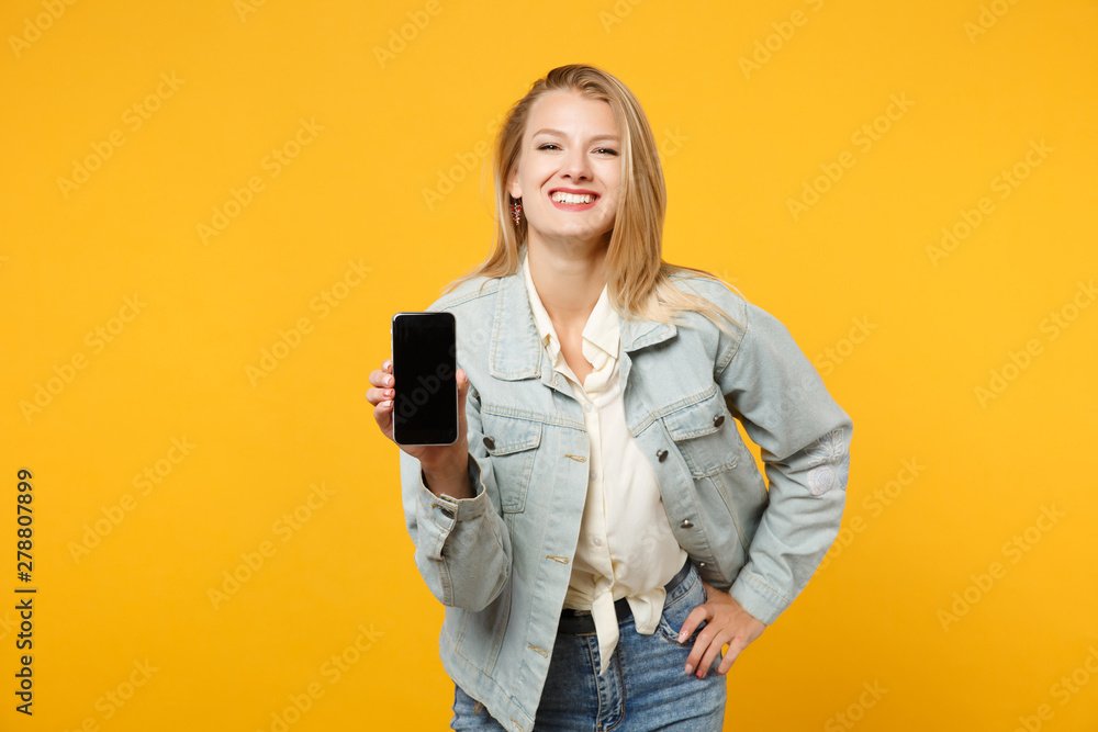 Portrait of cheerful young woman in denim casual clothes holding mobile phone with blank empty screen isolated on yellow orange wall background in studio. People lifestyle concept. Mock up copy space. Portrait of cheerful young woman in denim casual clothes holding mobile phone with blank empty screen isolated on yellow orange wall background in studio. People lifestyle concept. Mock up copy space.