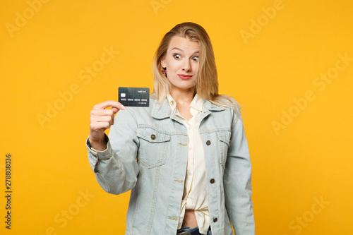 Portrait of excited perplexed young woman in denim casual clothes holding credit bank card isolated onbright yellow orange wall background in studio. People lifestyle concept. Mock up copy space.