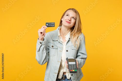 Pensive pretty young woman holding wireless modern bank payment terminal to process and acquire credit card payments isolated on yellow orange background. People lifestyle concept. Mock up copy space.
