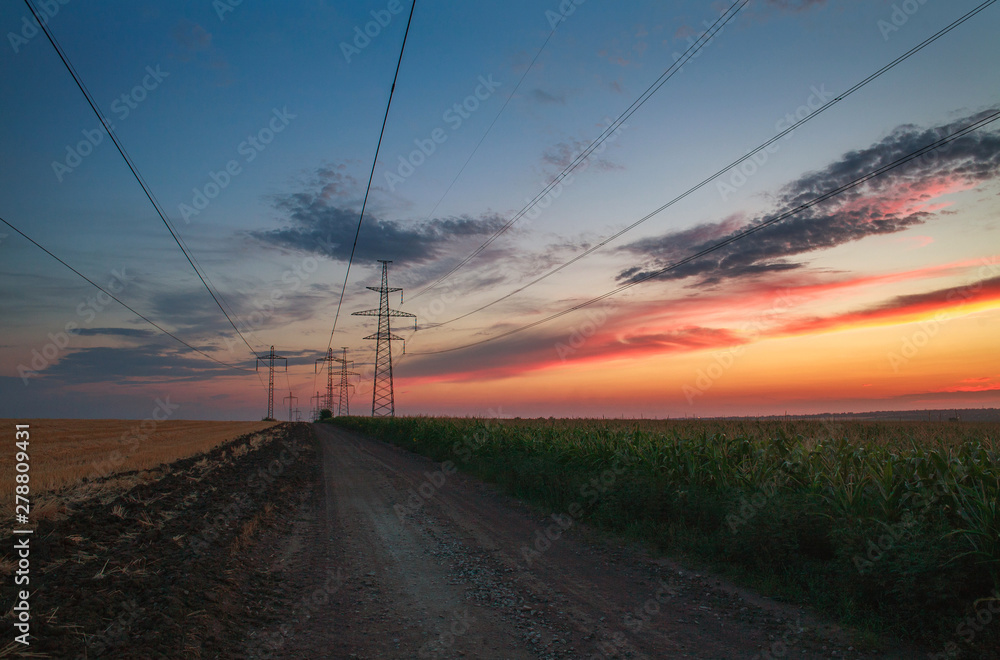Electrical power pylons on road in agricultural fields at sunset sky background