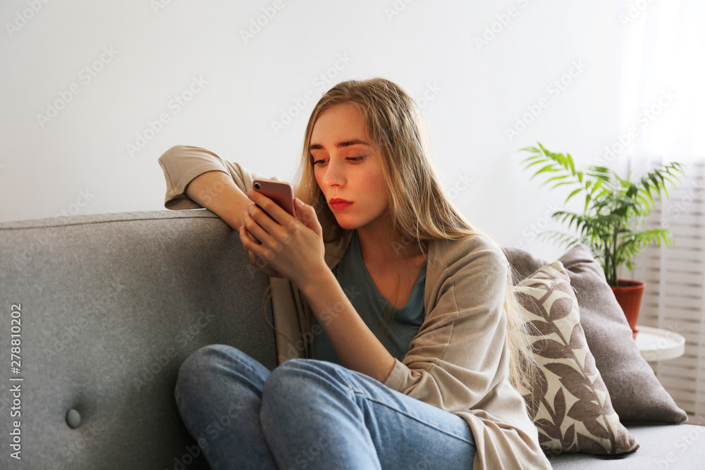 Φωτογραφία Portrait of beautiful young woman with depressed facial expression sitting on grey textile couch holding her phone