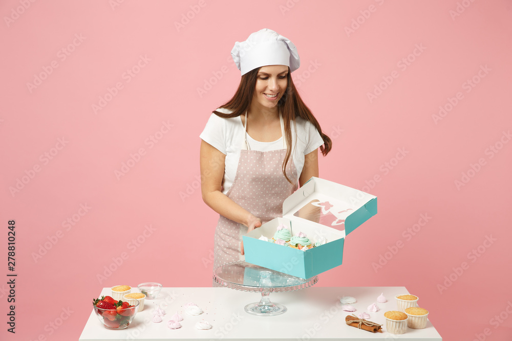 Housewife female chef cook confectioner or baker in apron white t-shirt, toque chefs hat packaging cake cupcake at table isolated on pink pastel background in studio. Mock up copy space food concept.